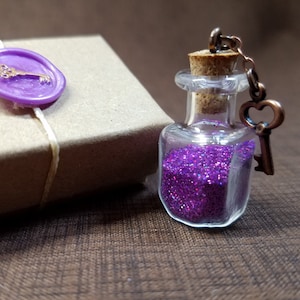 May include: A small glass bottle filled with purple glitter, attached to a key charm with a chain. The bottle is sitting on a brown surface.