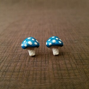 May include: A pair of blue and white mushroom-shaped earrings. The mushrooms have white polka dots on a blue background.