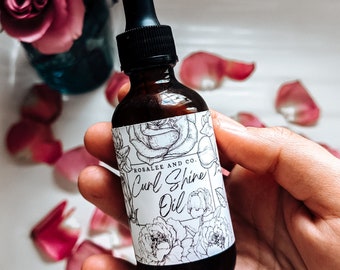 Puede incluir: Un frasco de vidrio ámbar oscuro con tapa de gotero negra y una etiqueta blanca con dibujos de líneas negras de rosas y el texto Rosalee and Co. Curl Shine Oil. El frasco se sostiene en la mano con pétalos de rosa rosados dispersos.