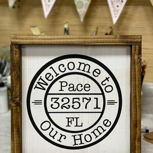May include: A wooden sign with a black circle and white text that reads "Welcome to Pace 32571 FL Our Home".