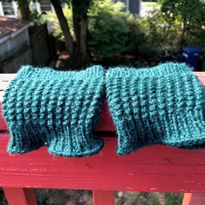 May include: Two teal green knitted wrist warmers, possibly made of wool, are laying on a red wooden railing. The warmers are textured and have a fuzzy appearance.