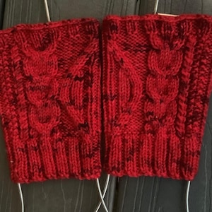 May include: A pair of red knitted wrist warmers with a cable knit design. The warmers are a vibrant red with flecks of darker red throughout. The cuffs are ribbed, and the main body features a textured pattern.