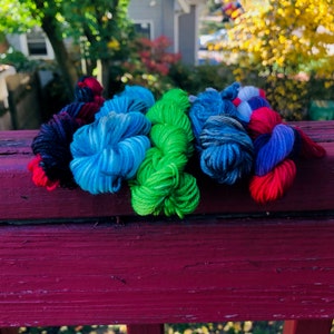 May include: Six skeins of yarn in various colors, including black, blue, green, red, and purple. The yarn is arranged in a row on a wooden surface.