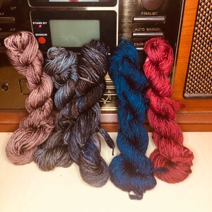May include: Four skeins of yarn in different colors: a dusty rose, a dark grey with blue flecks, a dark blue with green flecks, and a deep red.