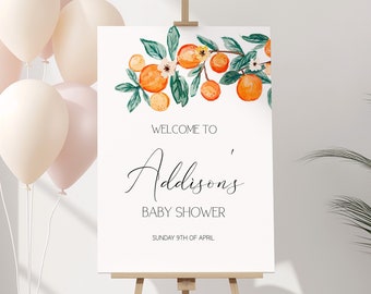 May include: A baby shower welcome sign on an easel. The sign is white with the text "Welcome to Addison's Baby Shower" and orange fruit and green leaf illustrations. A bundle of peach-colored balloons is to the left.