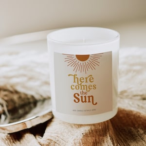 Puede incluir: Vela blanca con una etiqueta que dice "Here comes the Sun" con un gráfico de sol en naranja y marrón. La vela está sobre una superficie de tela beige.