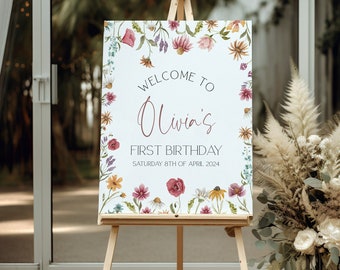 May include: A white welcome sign with a floral border and the text "Welcome to Olivia's First Birthday Saturday 8th of April 2024". The sign is on an easel in front of a vase of dried flowers.