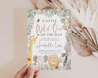 Può includere: Un invito a una festa di baby shower bianco con tema giungla. L'invito presenta una giraffa, un elefante, un leone e una zebra con foglie e fiori verdi. Il testo recita "A Little Wild One Is On The Way" e include il nome "Annabelle Lewis", la data e l'ora della festa, e l'indirizzo.