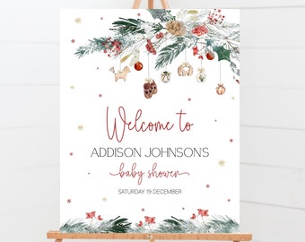 Può includere: Un cartello di benvenuto per baby shower bianco su un cavalletto di legno. Il cartello presenta un design a tema natalizio con rami sempreverdi, bacche rosse e ornamenti appesi. Il testo recita "Welcome to Addison Johnson's baby shower" e la data.