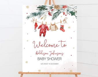 May include: A white sign on an easel with a Christmas baby shower theme. The sign reads "Welcome to Addison Johnson's BABY SHOWER SATURDAY 19 DECEMBER". The top of the sign features a garland of pine boughs and baby clothes hanging on a clothesline.