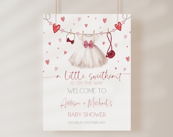 May include: A pink and white baby shower invitation with a watercolor illustration of a white dress, a pink bow, and a pink purse hanging on a clothesline with red hearts. The text reads "A Little Sweetheart is on the way. Welcome to Addison + Michael's Baby Shower. Saturday 11th February."