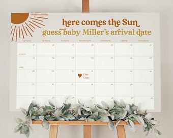 May include: A white calendar with the text "here comes the Sun guess baby Miller's arrival date" in gold lettering. A sun graphic is in the upper left corner. The calendar has a due date marked with a heart. A wooden easel supports the calendar.