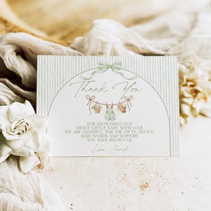 May include: A baby shower thank you card with a green bow and a clothesline with baby clothes illustrations. The card has a striped background and the words "Thank You" in script. The text reads "For showering our sweet little baby with love..." and is signed "Love, Sarah."