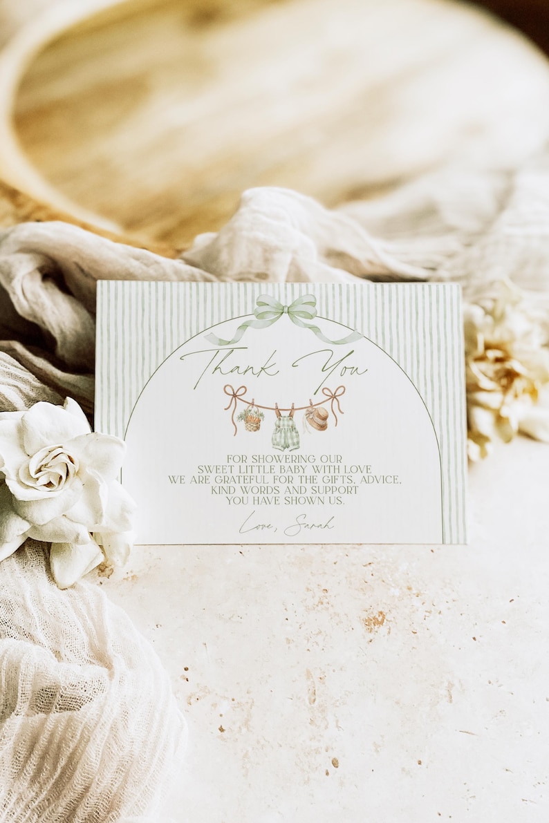 May include: A baby shower thank you card with a green bow and a clothesline with baby clothes illustrations. The card has a striped background and the words "Thank You" in script. The text reads "For showering our sweet little baby with love..." and is signed "Love, Sarah."