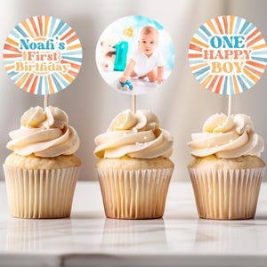 May include: Three cupcakes with white frosting and decorative toppers. One topper reads "Noafi's First Birthday" in blue, orange, and yellow. Another features a baby photo. The third says "ONE HAPPY BOY" in similar colors. The cupcakes sit on a white surface.