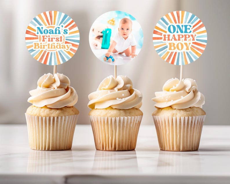 May include: Three cupcakes with white frosting and decorative toppers. One topper reads "Noafi's First Birthday" in blue, orange, and yellow. Another features a baby photo. The third says "ONE HAPPY BOY" in similar colors. The cupcakes sit on a white surface.