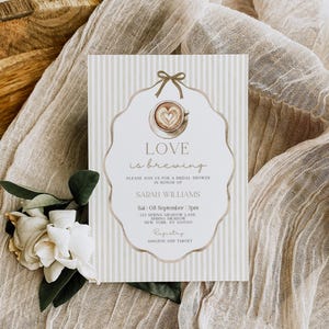 May include: Bridal shower invitation with a coffee cup design and the words "Love is brewing." The invitation is on a striped background with the event details. A white flower and beige fabric are in the background.