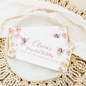 May include: A white chocolate bar wrapper with a pink floral design and a fairy illustration. The text on the wrapper reads "Olivia's fairy first birthday. Thank you for celebrating with me."