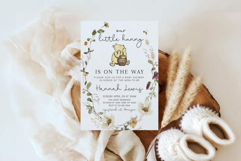 May include: A Winnie the Pooh themed baby shower invitation with a floral wreath and a bee. The text reads "our little hunny is on the way".