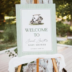 Pode incluir: Uma placa de boas-vindas para um chá de bebê, com uma ilustração de um carrinho de golfe. A placa tem um fundo listrado verde e branco com as palavras "WELCOME TO Sarah Smith's BABY SHOWER" e a data "SATURDAY APRIL 08". A placa é exibida em um cavalete de madeira.