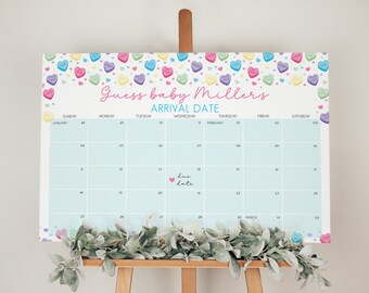 Op de afbeelding: Een lichtblauwe kalender met de tekst "Guess baby Miller's ARRIVAL DATE". De kalender is versierd met kleurrijke hartvormige snoepjes en een slinger van groene bladeren. De kalender wordt weergegeven op een houten ezel.