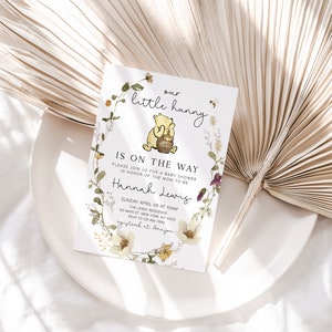 May include: A Winnie the Pooh themed baby shower invitation with a floral wreath and a cartoon image of Winnie the Pooh holding a honey pot. The text reads "our little hunny is on the way" and includes the name "Hannah Lewis" and details for the baby shower.