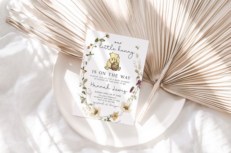 May include: A Winnie the Pooh themed baby shower invitation with a floral wreath and a cartoon image of Winnie the Pooh holding a honey pot. The text reads "our little hunny is on the way" and includes the name "Hannah Lewis" and details for the baby shower.