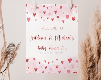 May include: A white sign with a pink and red watercolor background with hearts. The text reads "Welcome to Addison & Michael's baby shower" with a heart symbol. The date is "Saturday 11th Feb at 1PM".