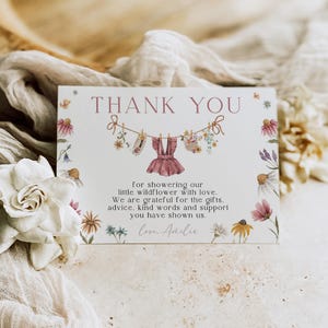 May include: A floral-themed thank you card with the words "THANK YOU" at the top. The card features a watercolor illustration of a baby dress and other baby clothes hanging on a clothesline. The text reads "for showering our little wildflower with love."