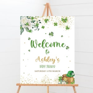 May include: Welcome to Ashley's Baby Shower sign with a gold pot of gold, shamrocks, and white flowers. The sign is on a white background with gold glitter.