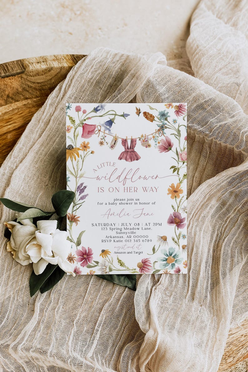 May include: A baby shower invitation with floral and watercolor design. The invitation features the text "A little wildflower is on her way" and details for the event. The invitation is surrounded by a floral border and rests on a beige fabric.