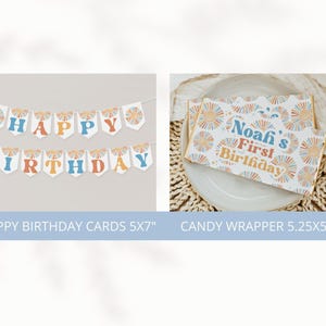 May include: A banner that says "HAPPY BIRTHDAY" in blue, orange, and yellow letters. The banner is strung with white string. A candy wrapper with the text "Noah's First Birthday" is also shown. The candy wrapper has a sunburst design.
