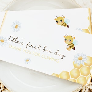 Puede incluir: Una tarjeta de agradecimiento blanca con un patrón de panal de abeja amarillo, dos abejas de dibujos animados y margaritas. El texto dice "Ella's first bee day. Thank you for coming."