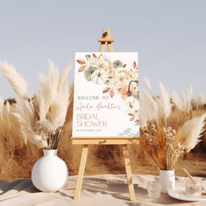 May include: A white sign with a watercolor illustration of fall flowers and pumpkins. The sign reads "Welcome to Amelia Hamilton's Bridal Shower on September 18, 2023".