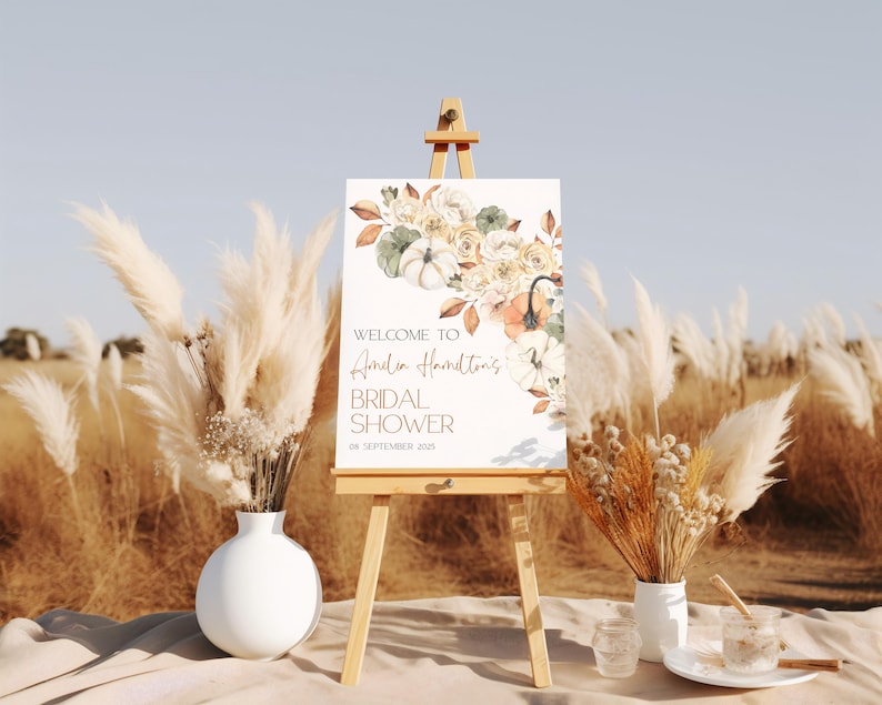 May include: A white sign with a watercolor illustration of fall flowers and pumpkins. The sign reads "Welcome to Amelia Hamilton's Bridal Shower on September 18, 2023".