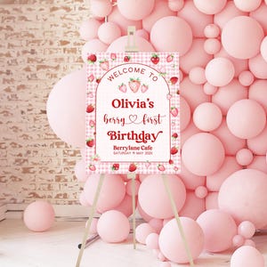 May include: Welcome to Olivia's berry first Birthday sign with a pink and white gingham background and red strawberries. The sign is on a wooden easel.