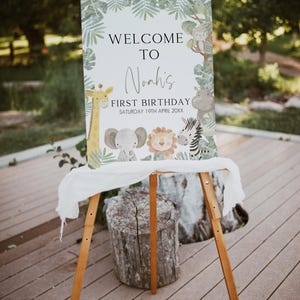 May include: Welcome to Noah's First Birthday sign with a jungle theme. The sign features a giraffe, elephant, lion, and zebra with green leaves and a white background.