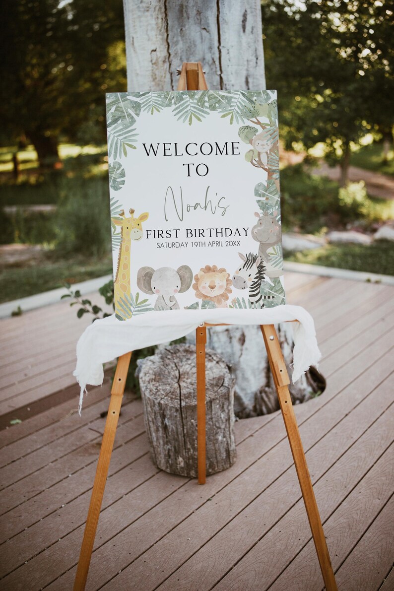 May include: Welcome to Noah's First Birthday sign with a jungle theme. The sign features a giraffe, elephant, lion, and zebra with green leaves and a white background.