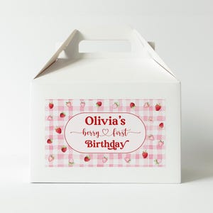 May include: White gable box with a handle, featuring a sticker with a pink gingham pattern and red strawberries. The sticker has the text "Olivia's berry first Birthday" in a red oval.