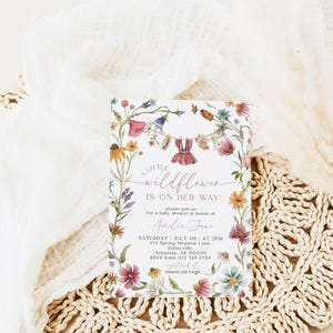 May include: A rectangular baby shower invitation card featuring a watercolor floral border with wildflowers and a clothesline displaying a small dress. The text reads: A LITTLE wildflower IS ON HER WAY, please join us for a baby shower in honor of Amelie Jane, SATURDAY JULY 08 AT 2PM, 123 Spring Meadow Lane, Sunnyville, Arkansas, AR 00000, RSVP Katie 012 345 6780, registered at Amazon and Target.