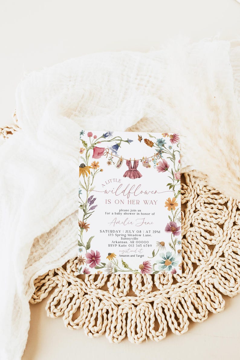 May include: A rectangular baby shower invitation card featuring a watercolor floral border with wildflowers and a clothesline displaying a small dress. The text reads: A LITTLE wildflower IS ON HER WAY, please join us for a baby shower in honor of Amelie Jane, SATURDAY JULY 08 AT 2PM, 123 Spring Meadow Lane, Sunnyville, Arkansas, AR 00000, RSVP Katie 012 345 6780, registered at Amazon and Target.