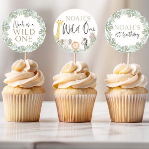 May include: Three cupcakes with white frosting and green and gold animal-themed cupcake toppers. The toppers say "Noah is a Wild One", "Noah's Wild One", and "Noah's 1st Birthday".