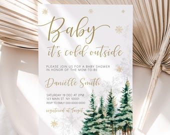 Op de afbeelding: Een witte babyborrel uitnodiging met gouden tekst die "Baby it's cold outside" zegt. De uitnodiging heeft een aquarel illustratie van een besneeuwd bos met dennenbomen.
