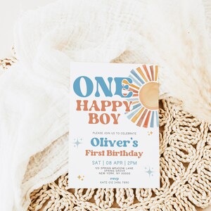 以下が含まれることがあります： 青とオレンジ色の「ONE HAPPY BOY」という文字が入った誕生日招待状。招待状には「Oliver's First Birthday」という文字とイベントの詳細が含まれています。招待状は織りのプレースマットの上にあります。