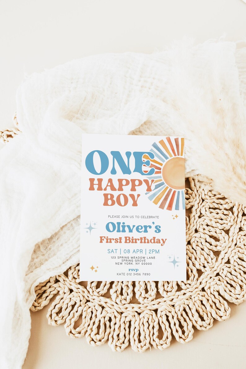 以下が含まれることがあります： 青とオレンジ色の「ONE HAPPY BOY」という文字が入った誕生日招待状。招待状には「Oliver's First Birthday」という文字とイベントの詳細が含まれています。招待状は織りのプレースマットの上にあります。