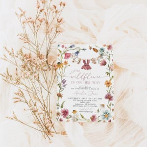 May include: A baby shower invitation with floral border and the text "A Little Wildflower Is On Her Way". The invitation features watercolor illustrations of flowers and baby clothes hanging on a clothesline. Dried flowers and fabric are in the background.