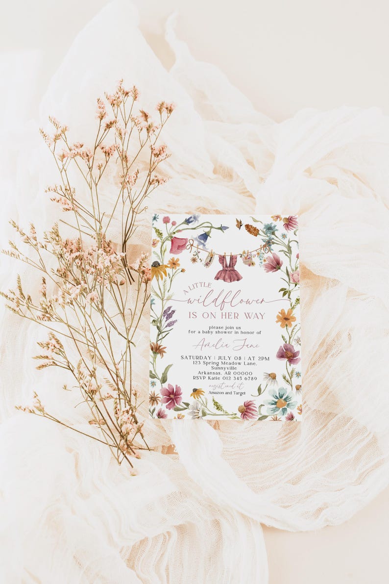 May include: A baby shower invitation with floral border and the text "A Little Wildflower Is On Her Way". The invitation features watercolor illustrations of flowers and baby clothes hanging on a clothesline. Dried flowers and fabric are in the background.