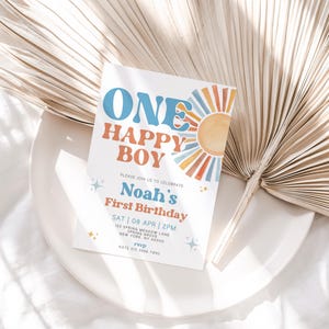 May include: A white invitation card with the text "ONE HAPPY BOY" in blue and orange, celebrating Noah's First Birthday. The card features a sun graphic and is placed on a white plate with a dried palm leaf.