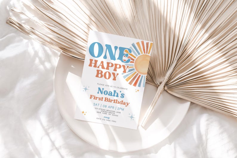 May include: A white invitation card with the text "ONE HAPPY BOY" in blue and orange, celebrating Noah's First Birthday. The card features a sun graphic and is placed on a white plate with a dried palm leaf.