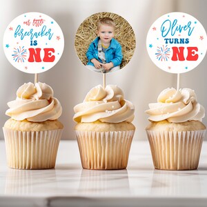 May include: Three cupcakes with white frosting and red and blue sprinkles topped with round paper cupcake toppers. The toppers feature a photo of a young boy, the text "our little firecracker is one" and the text "Oliver turns one".
